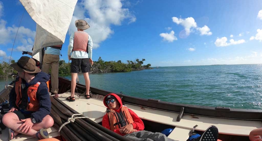 three people wearing life jackets stand and sit on a sailboat floating on calm blue water